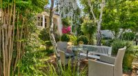 A cozy and intimate garden nook at Parker Cottage, featuring wicker chairs and a table set for tea, nestled among lush plants and shaded by mature trees.